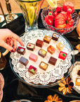 Assorted small desserts on a glass plate with a hand reaching for one, surrounded by decorative items.