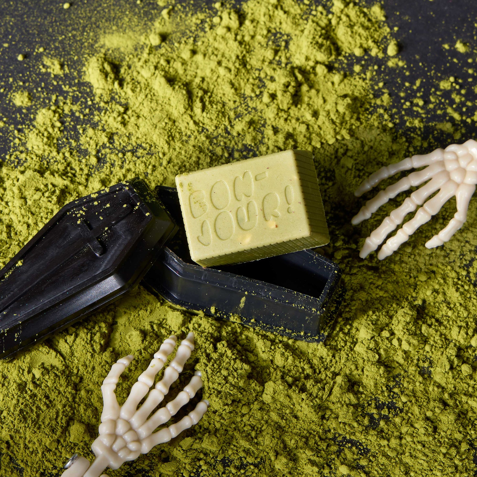 Matcha Crunch solid Bon on top of a black coffin, with matcha powder and skeleton hands in the background