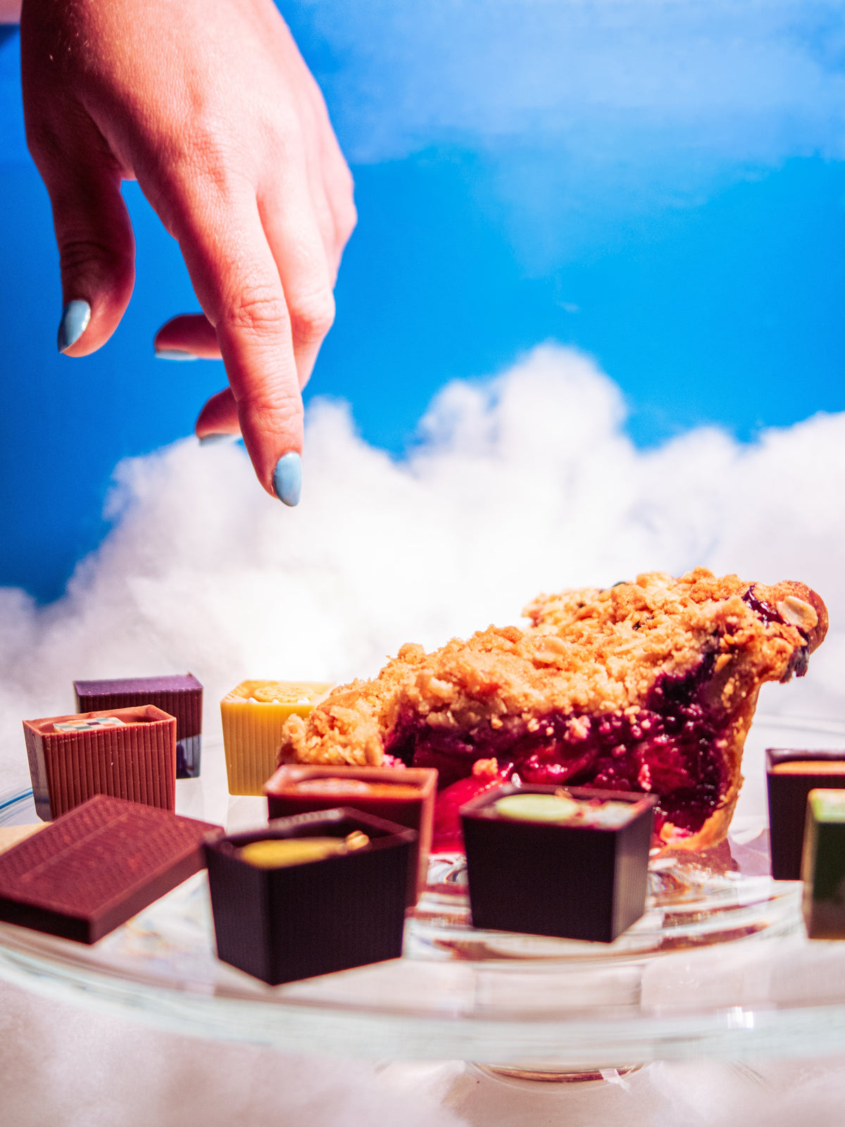 Glass table with Bons and a slice of pie on it. Hand from above reaching for the plate. Background of a blue sky with fluffy clouds