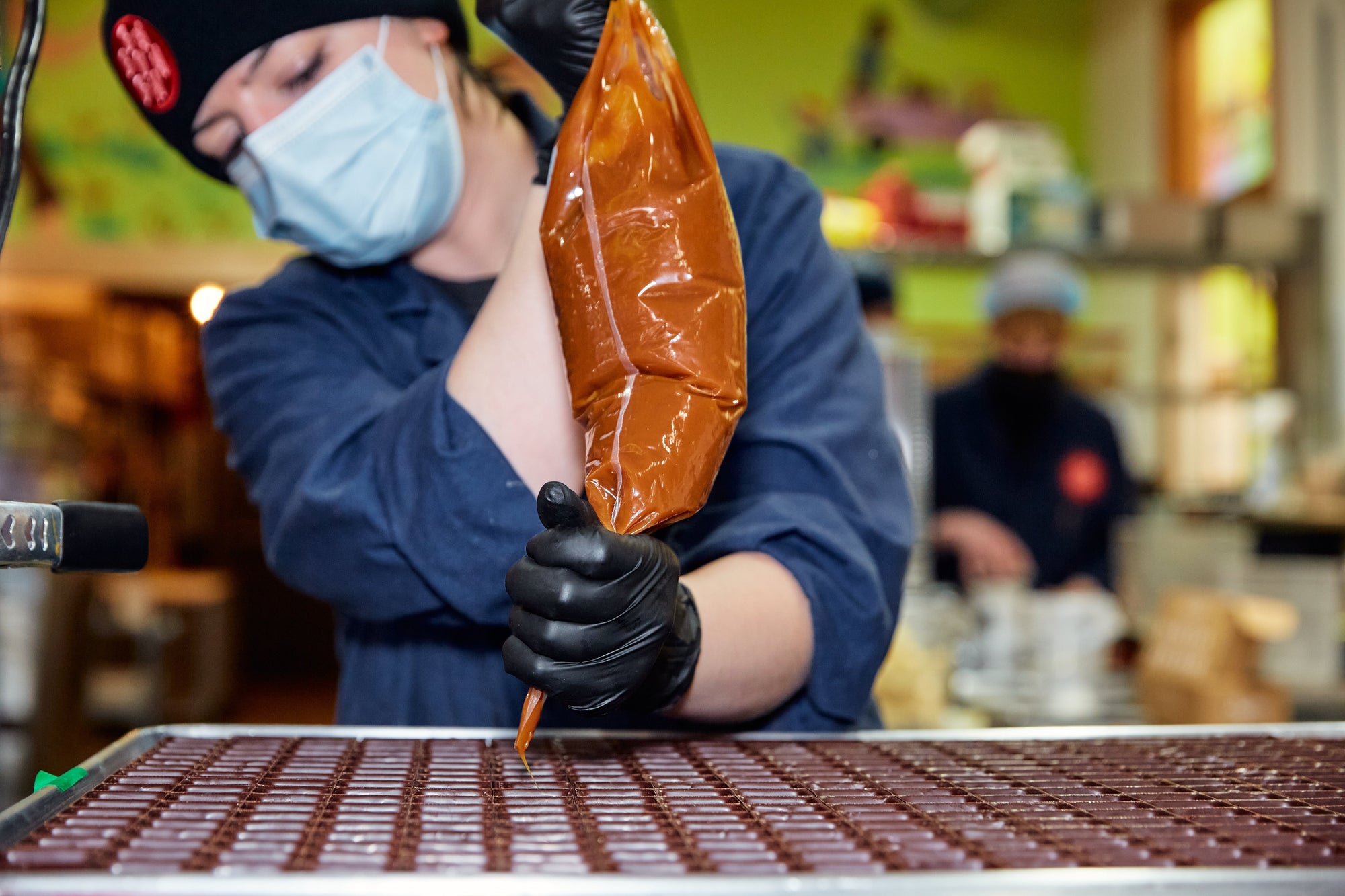 Woman piping ganache into empty chocolate shells with blurred background.