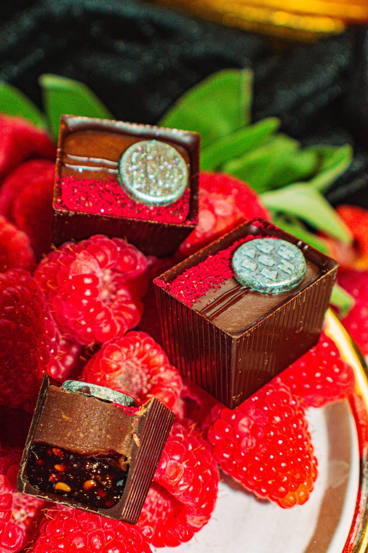 Baaa Baaa Boom Bon Bon Bon — a dark chocolate bon bon filled with goat’s milk caramel ganache, raspberry confiture, and candied sage for Bon Bon Bon's 2026 Valentine's Day Proper Fancy Collection. The bon bons are pictured on top of a bed of raspberries and sage in a fancy gold dish.