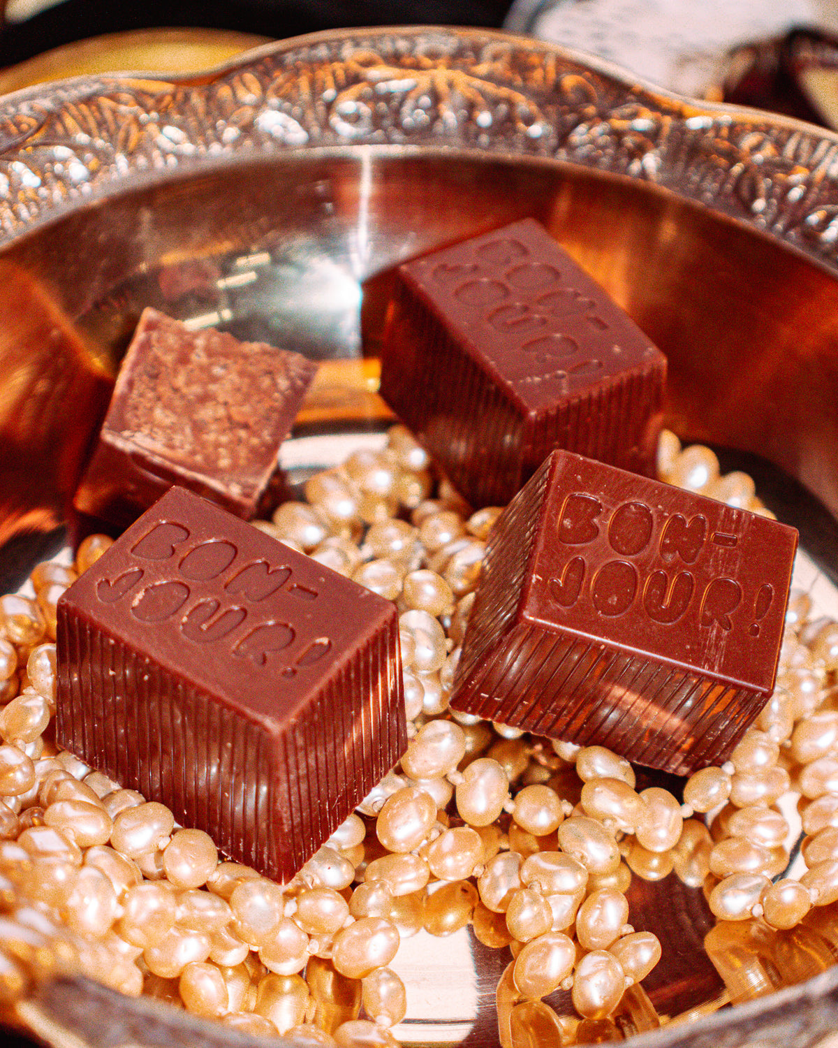 Buck Naked Bon Bon Bon — a milk and dark chocolate bon bon filled with buckweat cookie for Bon Bon Bon's 2026 Valentine's Day Proper Fancy Collection. The bon bon features 'Bon Jour!' text and is pictured on a bed of pearls in a decorative dish.