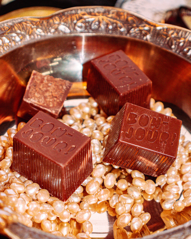 Buck Naked Bon Bon Bon — a milk and dark chocolate bon bon filled with buckweat cookie for Bon Bon Bon's 2026 Valentine's Day Proper Fancy Collection. The bon bon features 'Bon Jour!' text and is pictured on a bed of pearls in a decorative dish.