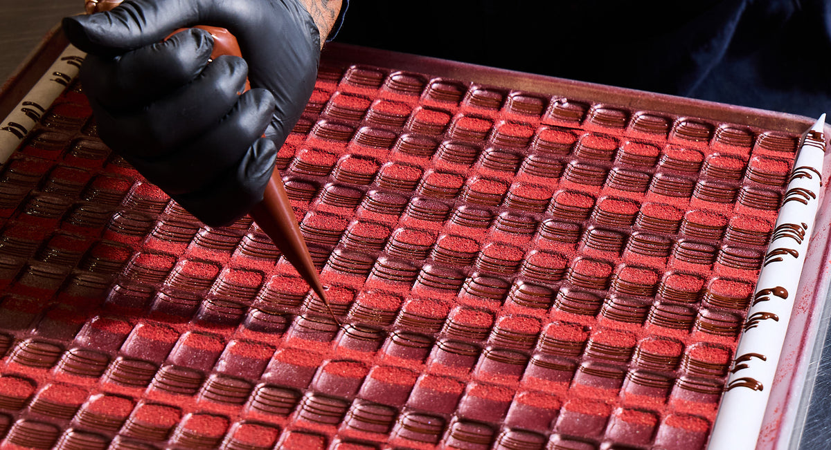 Hand wearing a black glove holding a piping bag over a tray of red bon bons