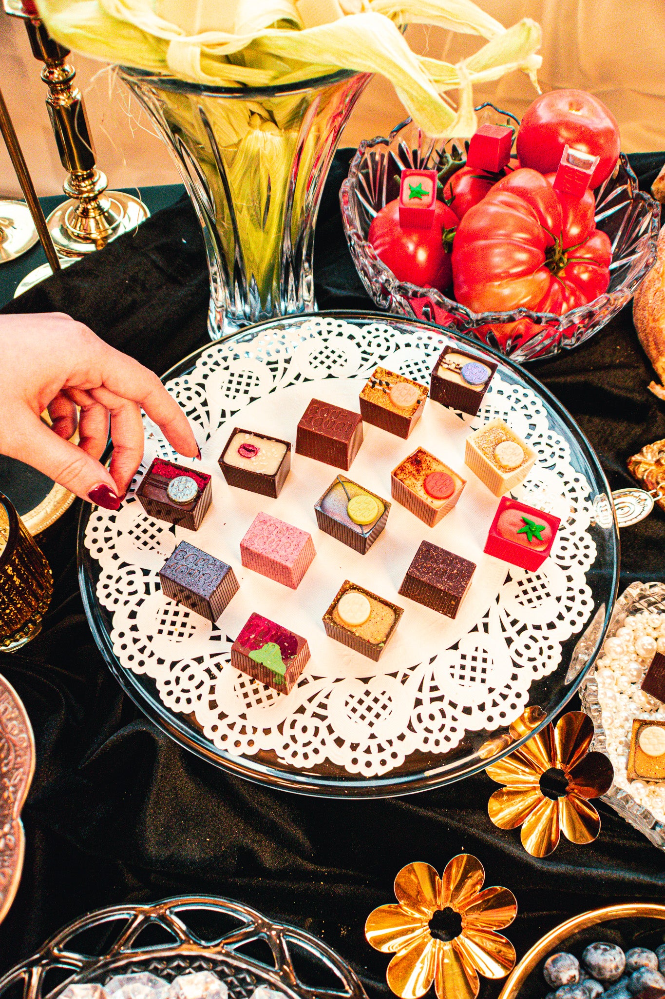 Assorted small desserts on a glass plate with a hand reaching for one, surrounded by decorative items.