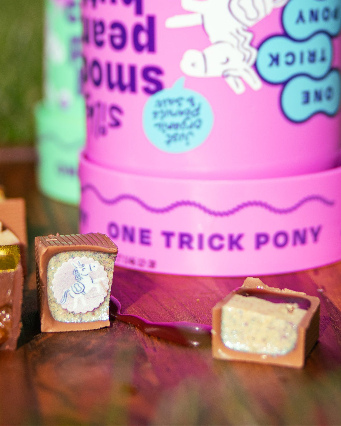 A pink tub of whipped peanut butter with a matching lid, labeled 'ONE TRICK PONY,' shown on a wooden surface outdoors with chocolate pieces in front.  chocolate with a small pony logo print. jelly on cuttingboard between bonbons