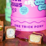 A pink tub of whipped peanut butter with a matching lid, labeled 'ONE TRICK PONY,' shown on a wooden surface outdoors with chocolate pieces in front.  chocolate with a small pony logo print. jelly on cuttingboard between bonbons