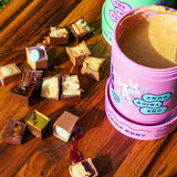 A pink tub of chocolate pudding labeled 'ONE TRICK PONY' sits on a wooden picnic table outdoors, with several chocolate pieces scattered around it and a mint green container behind it.
