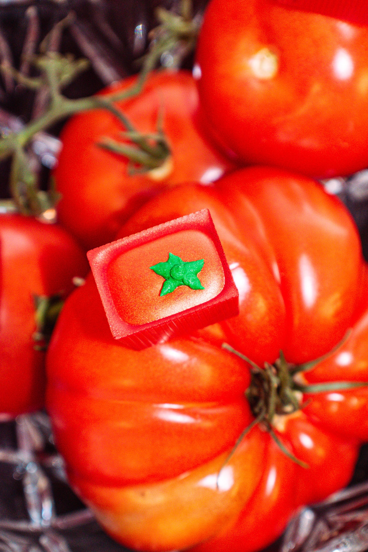 Head To-ma-toes Bon Bon Bon — a white chocolate bon bon filled with carmelized tomato tart tatin for Bon Bon Bon's 2026 Valentine's Day Proper Fancy Collection. The bon bon is decorated to look like a tomato and it is pictured sitting on top of heirloom tomatoes in a fancy glass dish.