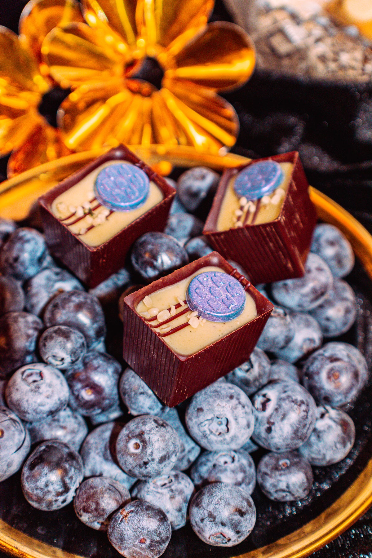Hemp Over Heels Bon Bon Bon — a dark chocolate bon bon filled with hemp seed gianduja, blueberry caramel, and hops infused ganache for Bon Bon Bon's 2026 Valentine's Day Proper Fancy Collection. The bon bons are pictured on top of a bed of blueberries in a fancy gold dish.