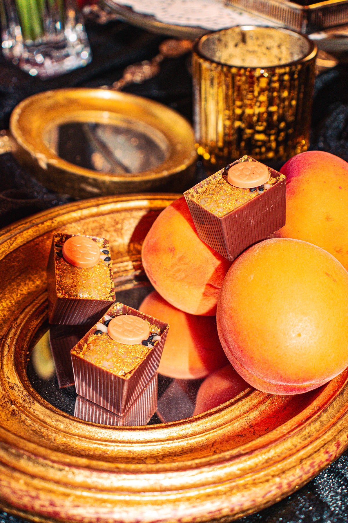 Lil’ Gold Mine Bon Bon Bon — a milk chocolate bon bon with a black sesame financier, apricot ganache, mascarpone, and toasted sesame for Bon Bon Bon's 2026 Valentine's Day Proper Fancy Collection. The bon bons are pictures alongside apricots on top of a fancy gold mirror.
