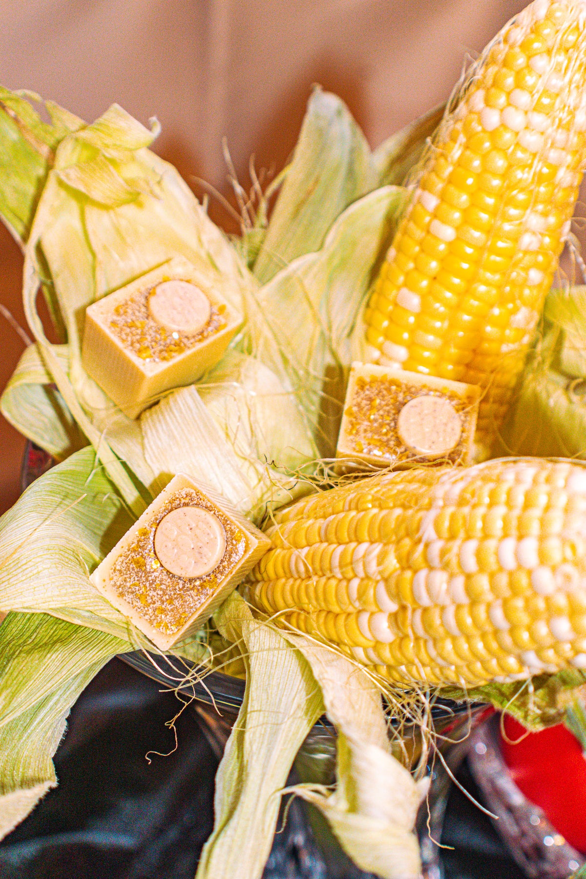 Might Sound Corny Bon Bon Bon — a white chocolate bon bon filled with a creamy coffee ganache and candied sweet corn for Bon Bon Bon's 2026 Valentine's Day Proper Fancy Collection. The bon bons are pictured amidst a bed of corn on the cob in a fancy glass vase.