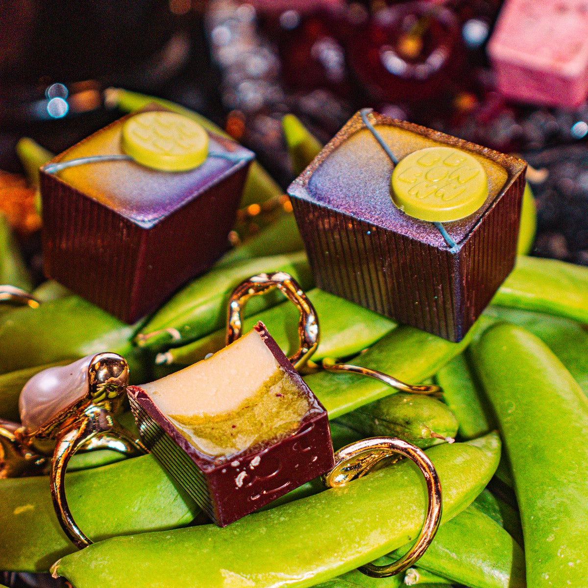 Peas Be Mine Bon Bon Bon — a dark chocolate bon bon with butter ganache, candied sweet peas, and butterfly pea flower for Bon Bon Bon's 2026 Valentine's Day Proper Fancy Collection. The bon bons are pictured on a bed of green beans with gold rings on them.