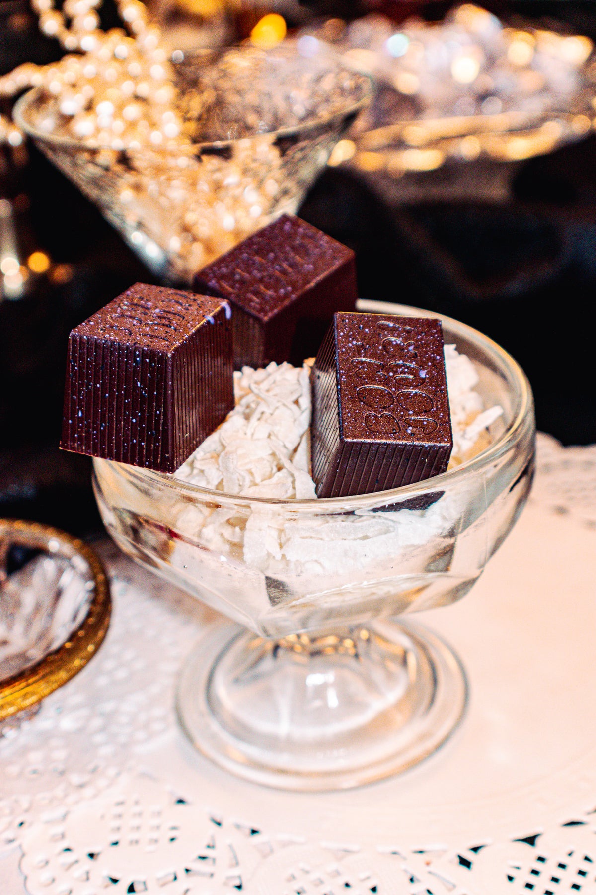 Saskatoon Macaro(o)n Bon Bon Bon — a dark chocolate bon bon with saskatoon confiture and lightly toasted coconut French macaron ganache for Bon Bon Bon's 2026 Valentine's Day Proper Fancy Collection. The bon bons are pictured on a bed of coconut in a fancy glass dish.