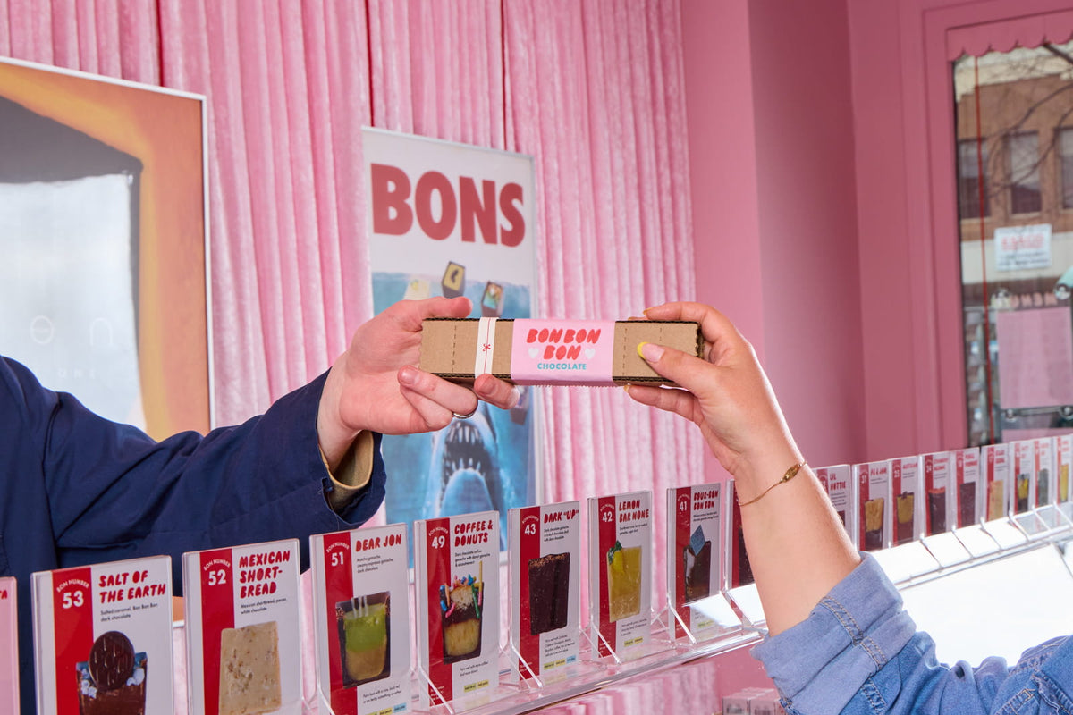 A retail staff member handing a box of Bons to a customer over the counter in Bon Bon Bon's shop in Birmingham, Michigan
