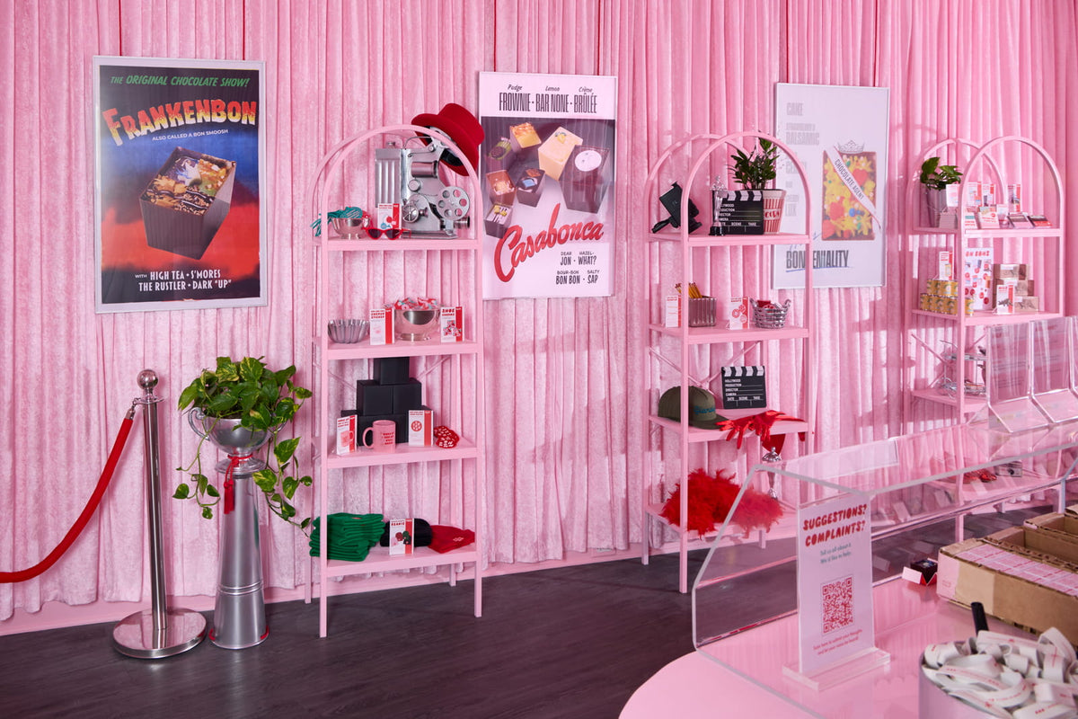 The interior of Bon Bon Bon's shop in Birmingham, Michigan, featuring a pink velvet curtain as a backdrop, pink shelving showcasing various Chocolate Oddities and merchandise, and Bon versions of classic Old Hollywood movie posters, like Frankenbon and Casabonca