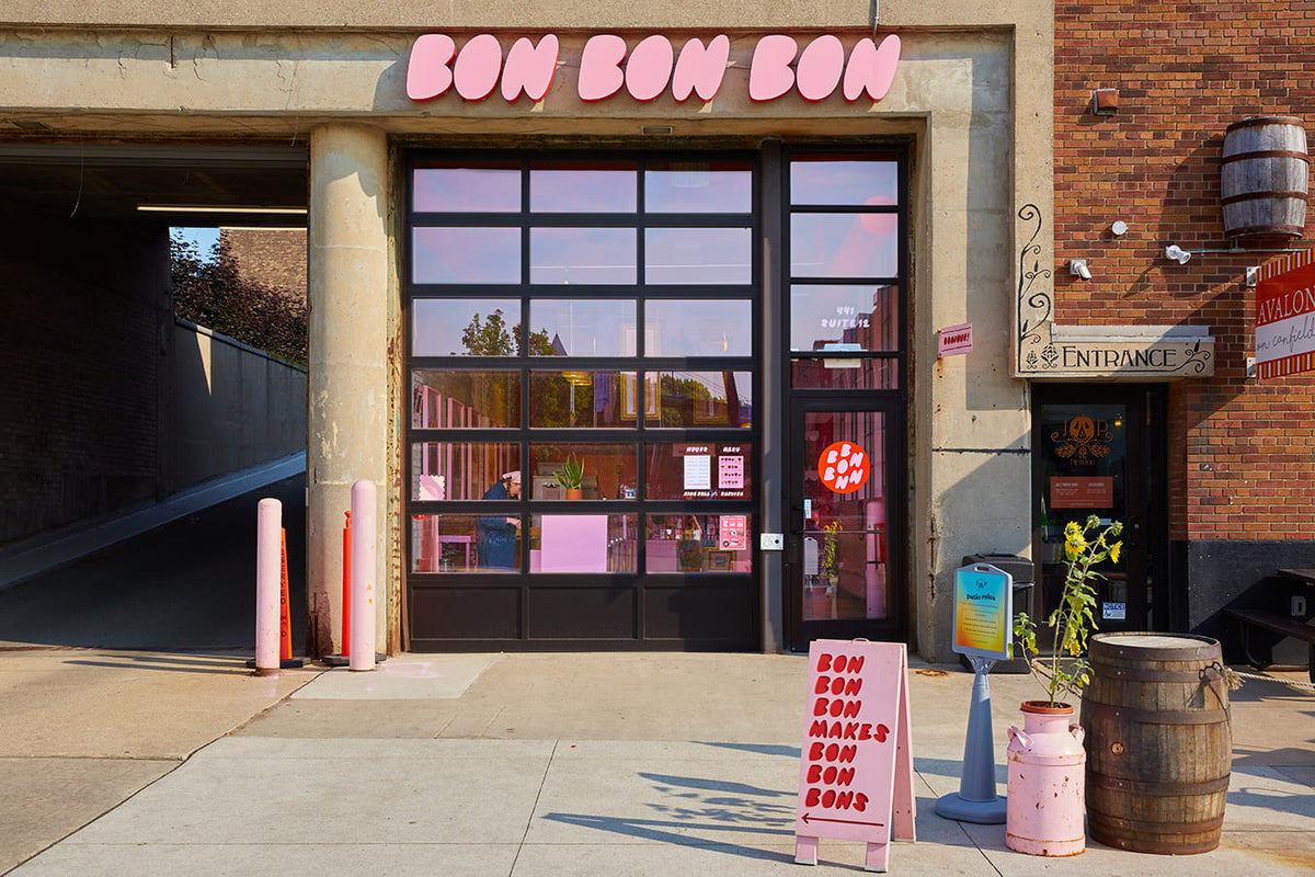 The exterior and front door of Bon Bon Bon's shop in Midtown Detroit. Out front, there is a pink sign with red text that reads "Bon Bon Bon makes bon bon bons", and the sign over the door features Bon Bon Bon's logo in their signature pink bubble-letter font