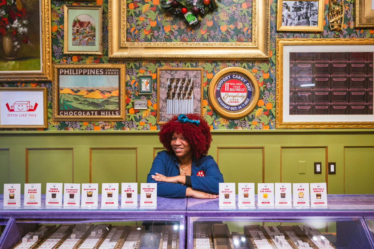 A retail employee standing behind the counter and greeting customers in Bon Bon Bon's shop in Parker's Alley in Detroit. Behind her is a gallery wall of framed art and chocolate-themed photography on a floral wallpapered wall.