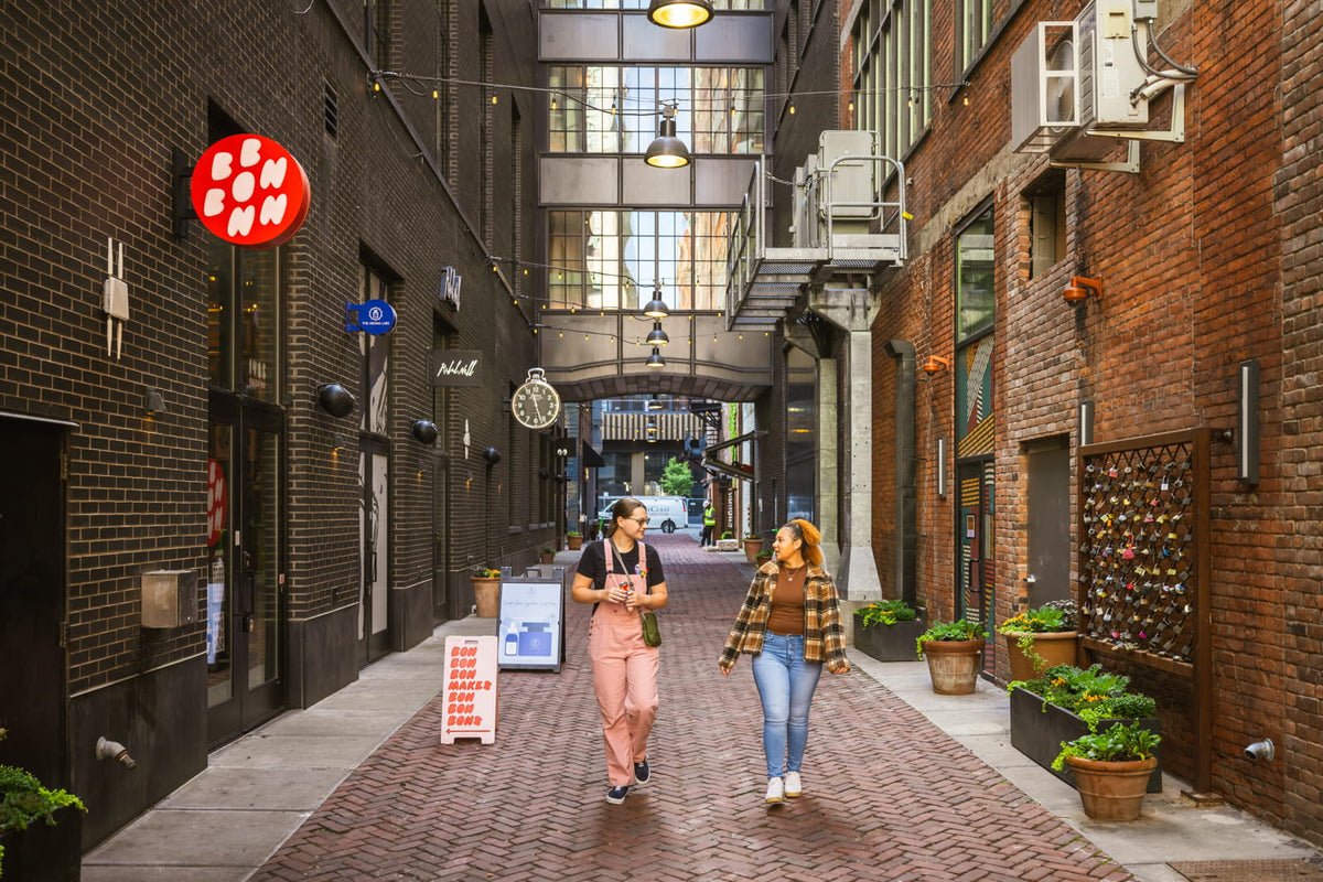 Two women walking through Parker's Alley in Detroit, passing Bon Bon Bon's shop on the left. There is a pink sign with red text reading "Bon Bon Bon makes bon bon bons" with an arrow pointing to the shop's door.